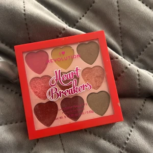 Heart Breakers ögonskuggspalett från Revolution - Säljer en färgglad ögonskuggspalett från Revolution med hjärtformade skuggor. Paletten innehåller åtta nyanser i rosa, grönt och guld, perfekt för att skapa kreativa looks.