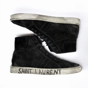 Saint Laurent höga svarta - FRI FRAKT PÅ THEARCHIVE.SE | Snygga svarta sneakers i mocka från Saint Laurent med handskriven logga på sulan. Skorna har en hög modell med snörning och en robust vit sula. Perfekta för en stilren look.                      Varumärke: Saint Laurant Storlek: EU 43 Skick: Nästan nyskick Material: Mocka Färg: Svart Anmärkningar: kommer utan låda Pris: 2699 SEK