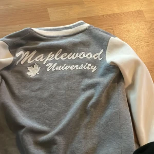 Grå och vit bomberjacka från Maplewood University - Säljer en grå och vit bomberjacka med tryck från Maplewood University. Jackan har långa ärmar och stängs med knappar framtill. Den har en ribbad krage och muddar, samt ett rött emblem på bröstet.