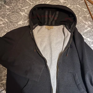 Mörkgrå hoodie från burberry  - Säljer en snygg mörkgrå hoodie från burberry  med dragkedja. Tröjan har praktiska fickor framtill och är perfekt för kyliga dagar. Den är tillverkad i ett mjukt material som ger en bekväm passform.
