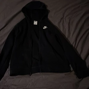 Svart huvtröja från Nike - Säljer en svart huvtröja från Nike med dragkedja och klassisk logga på bröstet. Perfekt för en avslappnad stil. Tröjan har långa ärmar och en luva. 