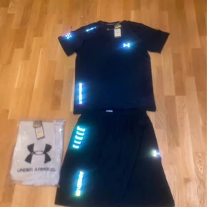 Svart t-shirt och shorts från Under Armour - Säljer en svart t-shirt och shorts från Under Armour med reflekterande detaljer. Perfekt för träning med sin lätta och bekväma passform. T-shirten har korta ärmar och är tillverkad i ett syntetmaterial som andas.