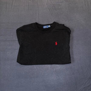 Svart Ralph Lauren T-shirt - Säljer en klassisk svart t-shirt från Ralph Lauren med det ikoniska röda logotypen på bröstet. T-shirten är kortärmad och gjord i mjukt bomullsmaterial. Perfekt för en stilren look. Har även använt få gånger!