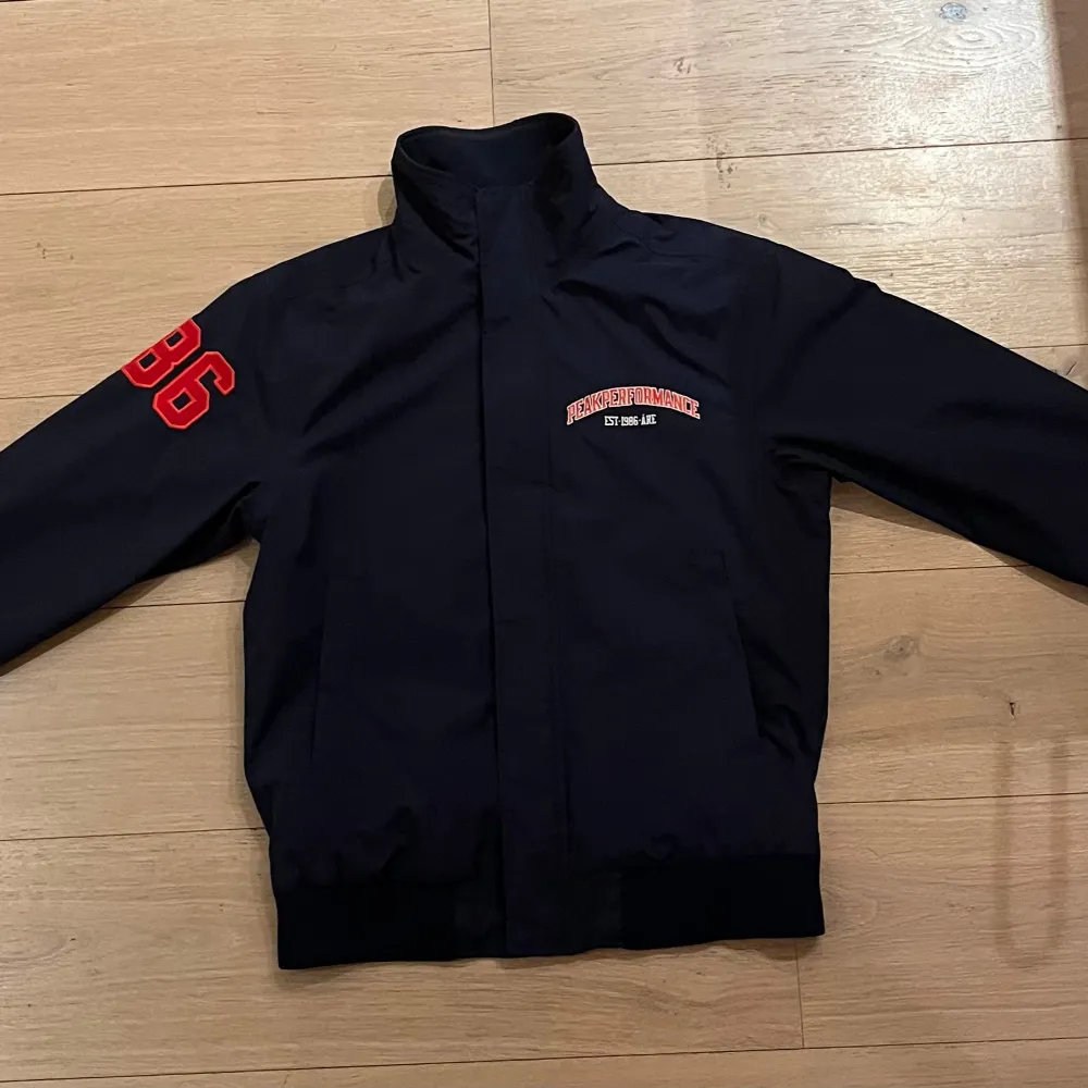 Säljer en mörk blå jacka från Peak Performance med röda detaljer. Jackan har en dragkedja framtill och är långärmad. Den har en broderad text på bröstet och siffran 86 på ärmen. Perfekt för en sportig look!. Takit.