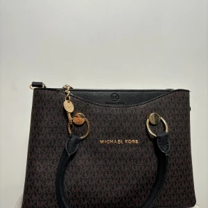Elegant och stilren Michael Kors-inspirerad väska – Perfekt för dig som älskar lyxiga detaljer!  - Letar du efter en snygg och funktionell väska som ger dig en lyxig känsla utan att kosta en förmögenhet? Då är den här väskan perfekt för dig! Den är i princip oanvänd (endast använd en gång) och ser helt ny ut. -  Klassisk design – Stilren brun och svart färgkombination med de ikoniska MK-detaljerna i guld. - Perfekt storlek – Rymlig men ändå smidig att bära, passar både till vardags och finare tillfällen. Smart inredning, Flera fack och dragkedjor för att hålla dina saker organiserade. Ej äkta
