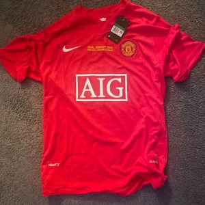 Röd fotbollströja från Nike - Säljer en röd fotbollströja från Nike med tryck för finalen i Moskva 2008. Tröjan har korta ärmar och AIG-logga på framsidan. På baksidan finns namnet 'Ronaldo' och nummer 7. Perfekt för fotbollsfans! Den är i xl men passar en L