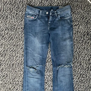 Blå jeans från Diesel - Snygga blå lågmidjadr jeans från Diesel med slitna detaljer på knäna. De har en klassisk femficksdesign och en knappgylf. 