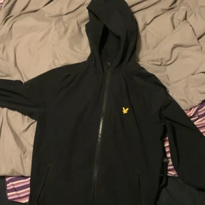 Svart vindjacka från Lyle & Scott - Säljer en svart huvtröja från Lyle & Scott med dragkedja och en gul logga på bröstet. Tröjan har långa ärmar och en praktisk huva. Perfekt för en avslappnad stil. Stor storlek passar mig som vanligtvis har s eller m. 