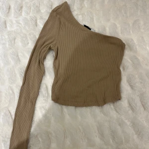 Beige oneshoulder topp från NA-KD - Snygg beige oneshoulder topp från NA-KD i ribbat material. Perfekt för en trendig look med sin unika design och långa ärm. Passar bra till både jeans och kjol.