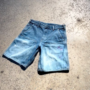 MARES ”MONEY PRINT” JORTS - Säljer ett par coola blå jeansshorts med unika färgstänk i lila och vitt. Shortsen har en klassisk femficksdesign och är perfekta för en avslappnad stil. De har en knapp- och dragkedjestängning framtill.