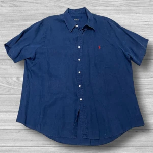 Blå kortärmad skjorta från Ralph Lauren - Snygg blå kortärmad skjorta från Ralph Lauren med klassisk krage och knappar framtill. Den har en liten röd broderad logga på bröstet som ger en stilren touch. Perfekt för en avslappnad men ändå stilfull look.