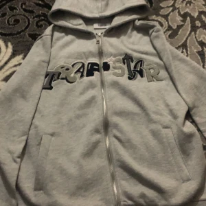 Grå hoodie från Trapstar - Säljer en grå hoodie från Trapstar med dragkedja och huva. Den har ett coolt broderat märke på framsidan i olika nyanser av grått och svart. Perfekt för en avslappnad stil.