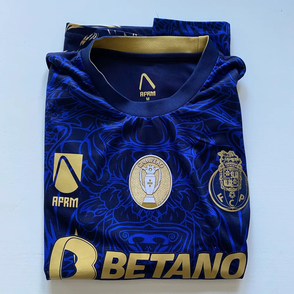 Snygg blå fotbollströja med ett coolt drakmotiv i guld. Tröjan har korta ärmar och flera logotyper på framsidan, inklusive Betano och Super Bock. Perfekt för fotbollsfans som vill sticka ut på läktaren!. T-paidat.