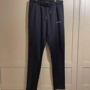 Mörkblå mjukisbyxor från Les Deux - Snygga mörkblå mjukisbyxor från Les Deux med dragsko i midjan och sidofickor. Perfekta för en avslappnad stil med en broderad logga på sidan. Byxorna har en bekväm passform och är tillverkade i ett mjukt material.