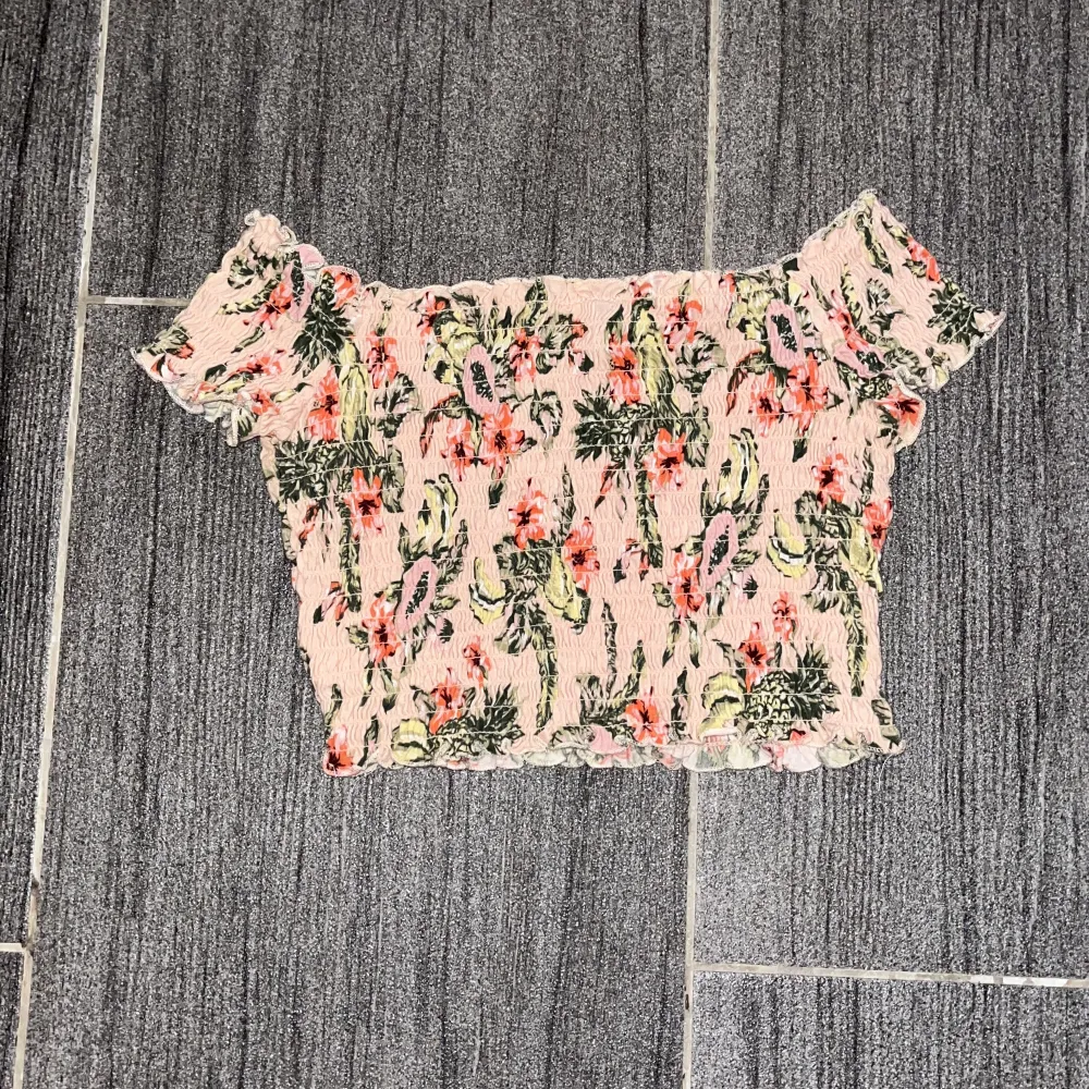 Säljer en superfin blommig offshoulder topp från Divided. Den är i en ljus färg med färgglada blommor och har en smockad, elastisk design som ger en bekväm passform. . Toppar.