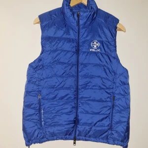 Blå dunväst från Ralph Lauren - Säljer en snygg blå dunväst från Ralph Lauren med RLX-logga på bröstet. Västen har en dragkedja framtill och två praktiska fickor med dragkedjor. Perfekt för kyliga dagar och ger en sportig look. Passar bra till både vardag och träning. Oanvänd utan prislapp. Storlek M