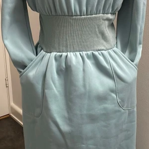 Ljusblå klänning med ribbad midja - ljusblåu klänning med en bred ribbad midja. Klänningen har långa ärmar och praktiska fickor framtill. Perfekt för vardag. Materialet känns mjukt och bekvämt mot huden.