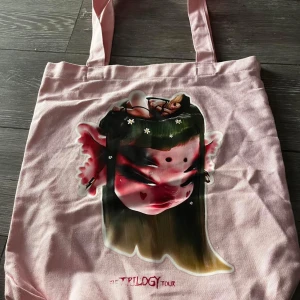 Rosa tygväska med konstnärligt tryck - Säljer en unik rosa tygväska med ett coolt konstnärligt tryck på framsidan. Perfekt för att bära med sig böcker eller använda som en shoppingväska. Väskan har två långa handtag och är i bra skick. Perfekt för vardagsbruk eller som en statement piece! Melanie Martinez vip bag
