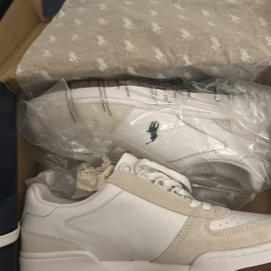 Vita sneakers från Polo Ralph Lauren - Snygga vita sneakers från Polo Ralph Lauren i nytt skick. De har en stilren design med vita och beige detaljer i skinn och mocka. Skorna har snörning och den klassiska Polo-loggan på sidan. Perfekta för både vardag och fest! Skorna var för små när jag köpte, de är helt nya och oanvända. Ny pris går för 1595kr, priset kan diskuteras!