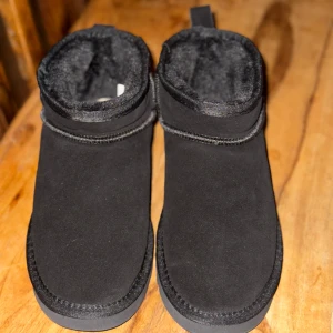 Svarta boots från UGG - Säljer ett par svarta UGG boots i mycket bra skick. De är gjorda av mocka och har en mjuk pälsfodring inuti för extra värme. Perfekta för kalla vinterdagar! Sulan är robust och ger bra grepp. Passar både till vardags och mysiga utflykter. Köpte dom nyligen men tyvärr så passar dom inte.