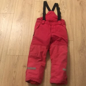 Rosa vinterbyxor med hängslen - Säljer ett par rosa vinterbyxor med justerbara svarta hängslen. Byxorna har reflexdetaljer vid benen och knappar framtill. Perfekta för kalla vinterdagar och utomhusaktiviteter. De är i bra skick och redo för nya äventyr!