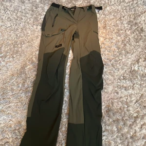 Gröna friluftsbyxor med dragkedjor - Säljer ett par gröna friluftsbyxor i bra skick. De har flera praktiska dragkedjor och förstärkta partier för extra hållbarhet. Perfekta för utomhusaktiviteter och äventyr i naturen. Passar bra för höst och vår.