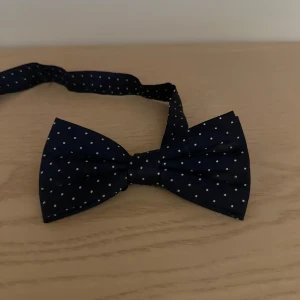 Mörkblå fluga med vita prickar - Snygg och stilren mörkblå fluga med små vita prickar. Perfekt för formella tillfällen eller när du vill lägga till en touch av elegans till din outfit. Flugan är justerbar för att passa de flesta halsstorlekar. Ett måste för garderoben!