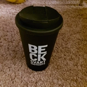 Svart termosmugg med text - Säljer en svart termosmugg med texten 'BECK SVART SNUTKAFFE' på ena sidan och 'C MORE' på den andra. Muggen har ett avtagbart lock med dricköppning. Perfekt för att hålla kaffet varmt på språng!
