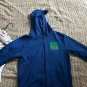 Blå hoodie från Polo Ralph Lauren - Säljer en snygg blå hoodie från Polo Ralph Lauren. Den har en dragkedja framtill och en stor logga i grönt och orange både fram och bak. Perfekt för en avslappnad stil och passar bra till både höst och vår. Den är i mycket bra skick och redo för nya äventyr!