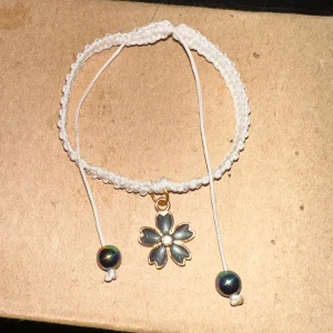Vitt armband med blomma och pärlor - Säljer ett superfint vitt armband med en söt blomma i guld och svarta detaljer. Armbandet har justerbara band med två skimrande pärlor i ändarna. Perfekt för att ge en bohemisk touch till din outfit!