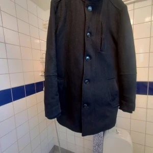 Svart ulljacka från Jack & Jones - Säljer en stilren svart ulljacka från Jack & Jones Premium. Jackan har en klassisk design med knappar framtill och en praktisk dragkedja på insidan för extra värme. Perfekt för höst och vinter med sin långa ärm ,eleganta look. Den är i behov kemtvätt