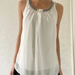 Vit ärmlös topp med dekorativ halsringning - Säljer en elegant vit ärmlös topp med en vacker dekorativ halsringning. Toppen är luftig och perfekt för varma dagar. Den har en lätt och flödande passform som ger en skön känsla. Perfekt för både vardag och festliga tillfällen!