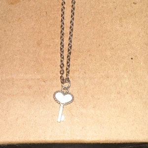 Silverfärgat halsband med hjärtformad nyckel - Säljer ett superfint silverfärgat halsband med en hängande nyckel i form av ett hjärta. Perfekt för att ge en romantisk touch till din outfit. Kedjan är enkel och stilren, vilket gör att den passar till alla tillfällen. Ett måste för alla smyckesälskare! 💖