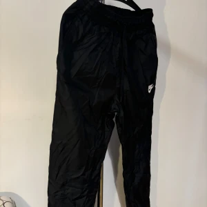 Svarta träningsbyxor från Nike - Säljer ett par svarta träningsbyxor från Nike i storlek S. Byxorna har en elastisk midja med snörning för bästa passform. Perfekta för träning eller en avslappnad dag. Nike-loggan är broderad på sidan. Sköna och stilrena! Helt nya!!  Nypris: 599kr