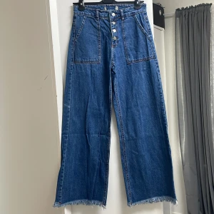 Blå jeans med hög midja och fransade kanter - Säljer ett par snygga blå jeans med hög midja och fransade kanter. De har en cool wide fit och knappar framtill. Perfekta för en avslappnad stil med en touch av edge. Passar både till vardags och fest!