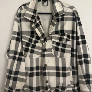 Rutig overshirt i svart och vit - Säljer en snygg rutig overshirt i svart och vit. Den har en avslappnad passform och är perfekt för lager-på-lager-stil. Jackan har knappar framtill och två stora bröstfickor med lock. Perfekt för höst och vår!