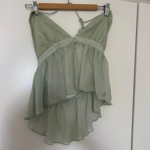 Grön ärmlös topp med volang - Säljer en superfin grön ärmlös topp med volangdetaljer. Toppen har en luftig och fladdrig design med knytband över halsen. Perfekt för sommardagar eller en festlig kväll. Den är i ett lätt och skönt material, med öppen rygg