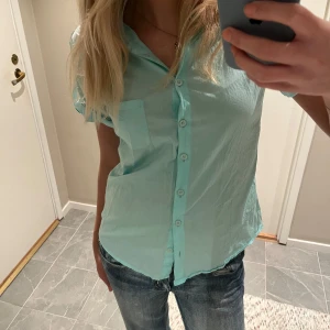 Ljusblå skjorta från Guess - Snygg ljusblå skjorta från Guess med korta ärmar och knappar framtill. Perfekt för en avslappnad stil. Skjortan har en klassisk krage och en bröstficka för extra stilpoäng. Även detaljer i ärmarna som gör skjortan lite mer speciell 