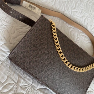 Brun midjeväska med kedjedetalj - Snygg brun midjeväska från Michael Kors med guldfärgad kedjedetalj. Väskan har ett justerbart midjeband och är perfekt för att bära dina nödvändigheter med stil. Den är tillverkad i ett slitstarkt material med ett diskret mönster.