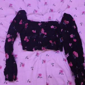 Svart blommig topp - Säljer en svart topp med rosa blommönster. Toppen har långa ärmar och en croppad design. Perfekt för en trendig look.