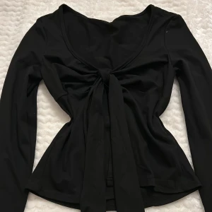 Svart långärmad topp med knyt - Snygg svart långärmad topp med knyt framtill. Toppen har en djup v-ringning och är perfekt för en stilren look. Materialet är mjukt och stretchigt för en bekväm passform. (oanvänd)