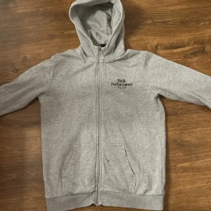 Grå hoodie från Peak Performance - Säljer en stilren grå hoodie från Peak Performance med dragkedja och huva. Tröjan har två praktiska fickor framtill och ett broderat märke på bröstet. Perfekt för en avslappnad look.