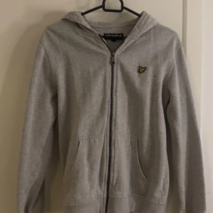 Grå hoodie från Lyle & Scott - Säljer en stilren grå hoodie från Lyle & Scott med dragkedja och huva. Tröjan har en broderad logotyp på bröstet och praktiska fickor framtill. Perfekt för en avslappnad stil.