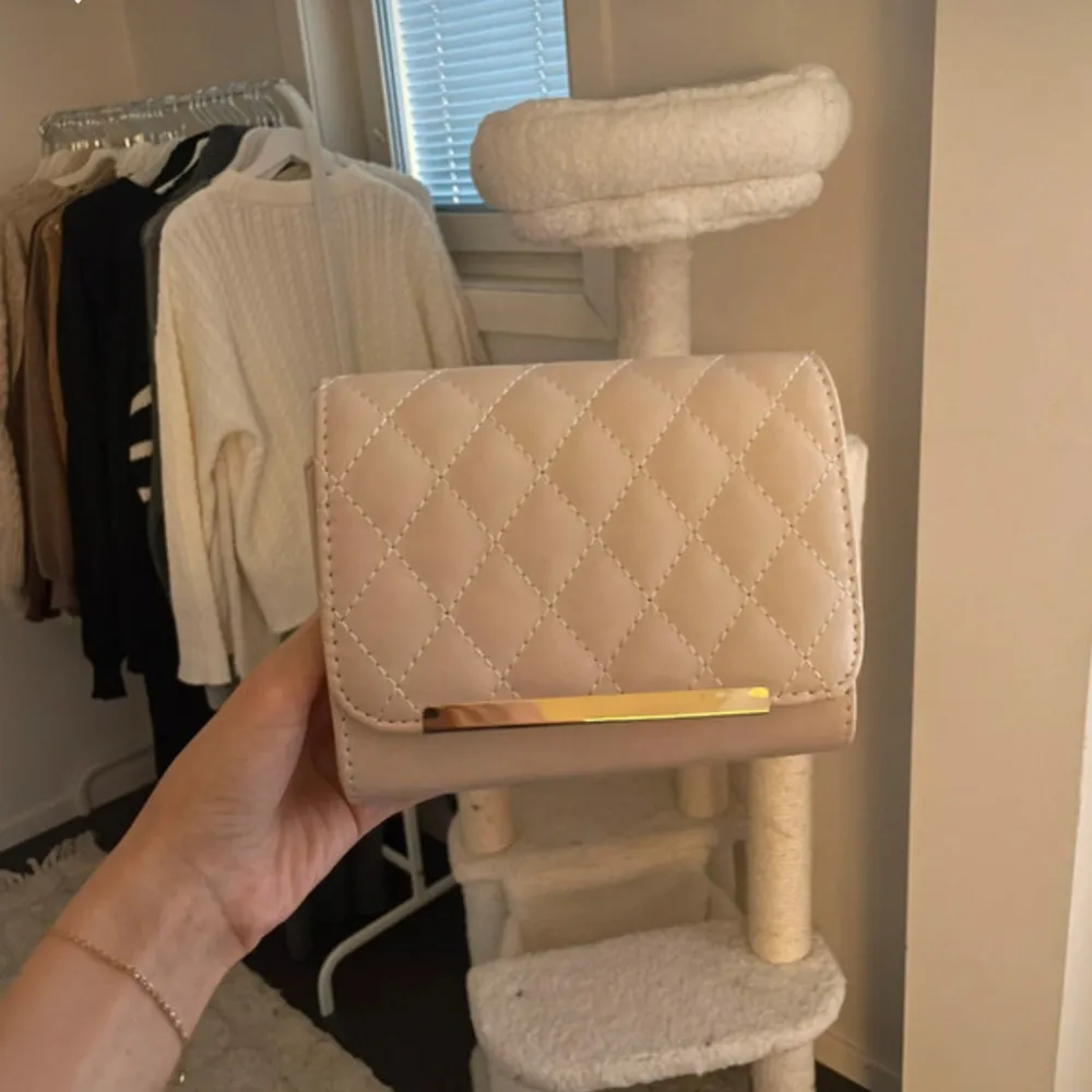 Säljer en beige handväska med quiltat mönster och en elegant gulddetalj på framsidan. Väskan har en praktisk dragkedja inuti för säker förvaring. Perfekt för att ge din outfit en chic touch. Finns något märke på baksidan men syns inte så mycket. . Laukut & Käsilaukut.