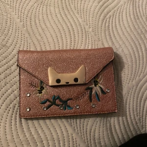 Rosa axelväska med kattdetalj - Säljer en söt rosa axelväska med en unik kattdetalj på framsidan. Väskan har broderade blommor och en kedja som axelrem. Perfekt för att ge en lekfull touch till din outfit.