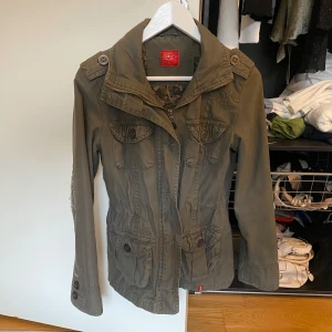 Olivgrön jacka från Meiyan - Snygg olivgrön jacka från Meiyan med coola broderade detaljer på ryggen och ärmarna. Jackan har en dragkedja och knappar framtill samt flera praktiska fickor. Insidan har ett camouflage-mönster som ger en extra edge. Perfekt för höst och vår!