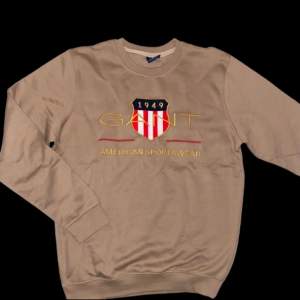 Säljer en beige sweatshirt från GANT med broderad logga och texten 'American Sportswear' på framsidan. Tröjan har långa ärmar och rund halsringning. Perfekt för en avslappnad stil. Har aldrig använts