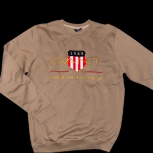 Beige sweatshirt från GANT - Säljer en beige sweatshirt från GANT med broderad logga och texten 'American Sportswear' på framsidan. Tröjan har långa ärmar och rund halsringning. Perfekt för en avslappnad stil. Har aldrig använts