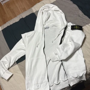 Vit hoodie från Stone Island - Säljer en äkta vit hoodie från Stone Island med dragkedja och huva. Den har en ikonisk patch på ärmen och ribbade muddar. Perfekt för en casual look.