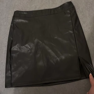 Svart kjol från Shein - Snygg svart kjol i skinnimitation från Shein. Kjolen har en stilren design med en liten slits på framsidan och dragkedja baktill. Perfekt för en trendig look.